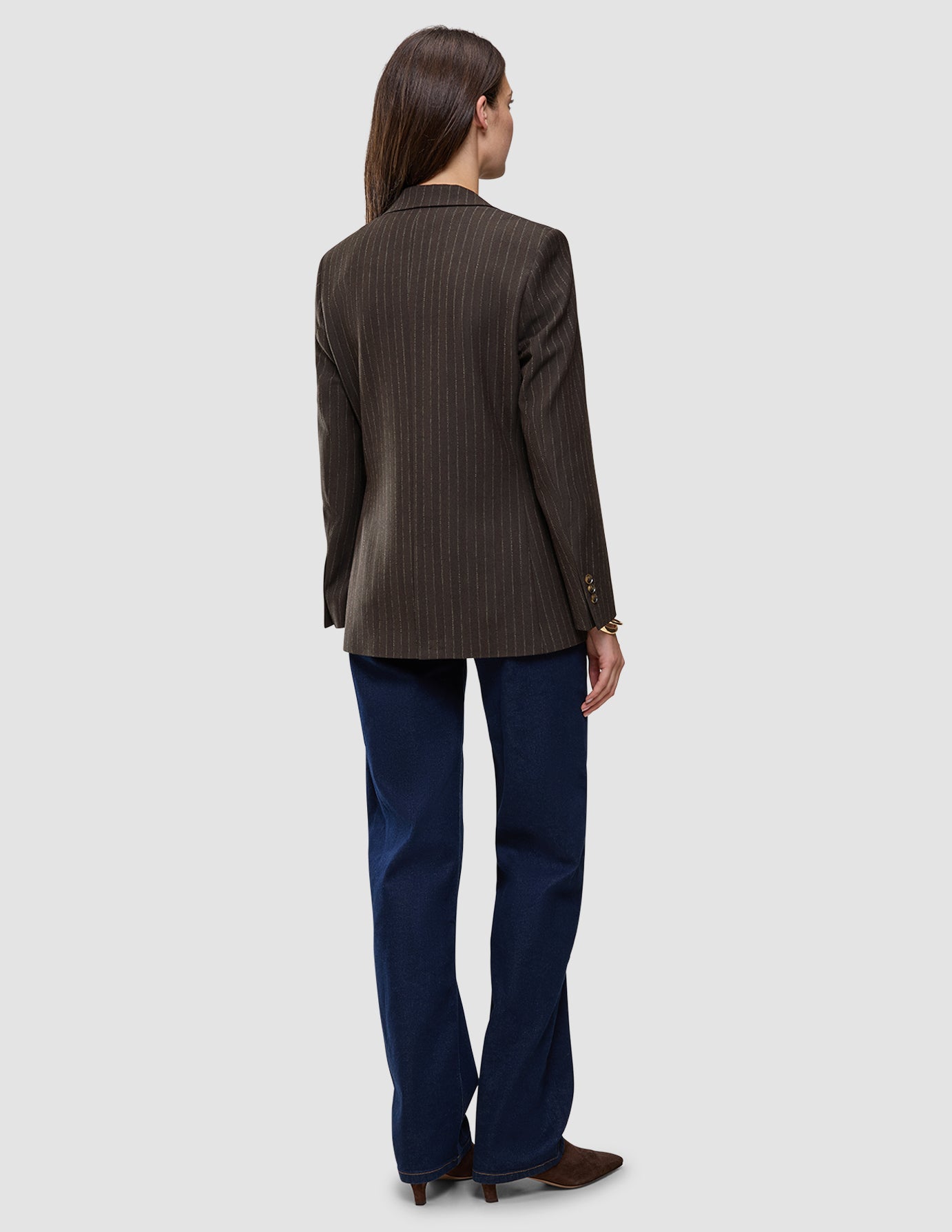 Eine Person, von hinten gesehen, die einen Women Serene Blazer Regular Walnut Herringbone, eine dunkelblaue Jeans und braune Schuhe trägt.
